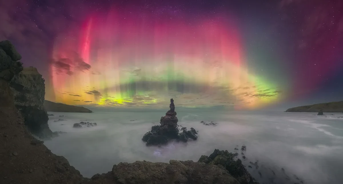 The aurora appears as a pink and yellow circle in the night sky. This image is taken from a rocky mountainside, with clouds filling the valley beneath